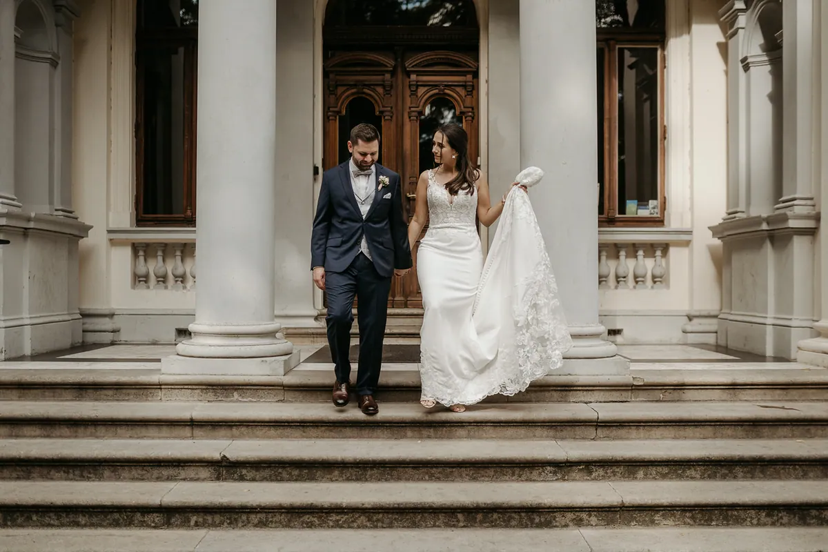 Jenny und Chris gehen Hand in Hand die Stufen einer Villa mit Säulen hinunter